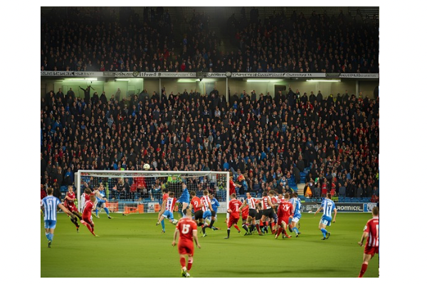 Action shot from the Huddersfield vs. Barnsley match, showing Huddersfield players crowding in on the opponents. The match ended 2-0 in favor of Huddersfield as fans in the background cheers on at the stadium.